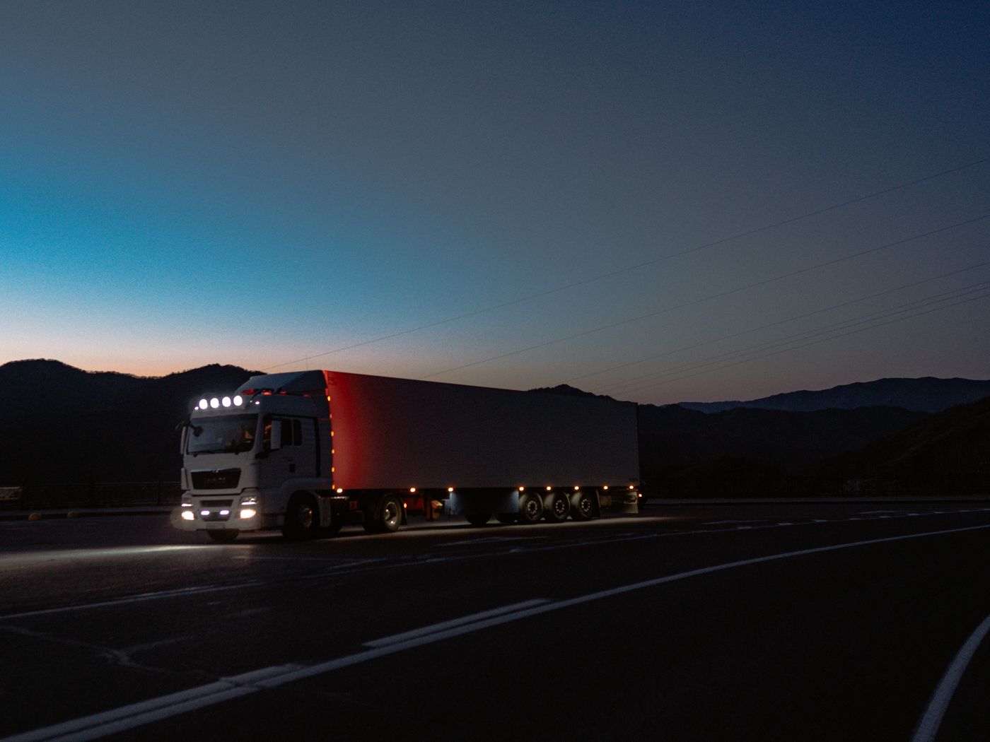 A white tractor-trailer with its lights on, parked on an empty highway at dusk with mountains behind