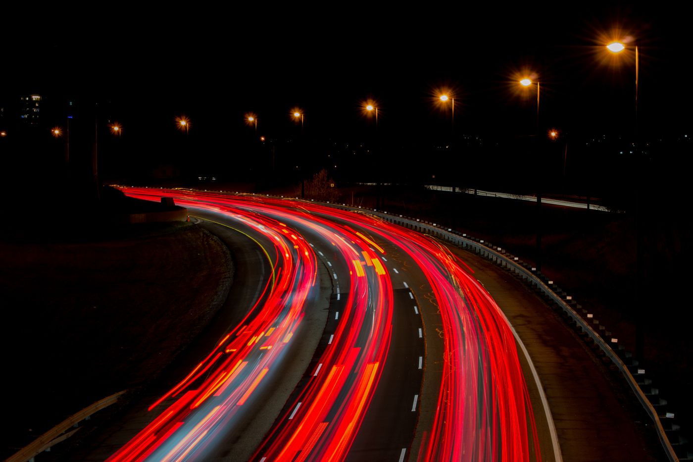 Red tail-light trails curving along a multi-lane highway at night under orange streetlights
