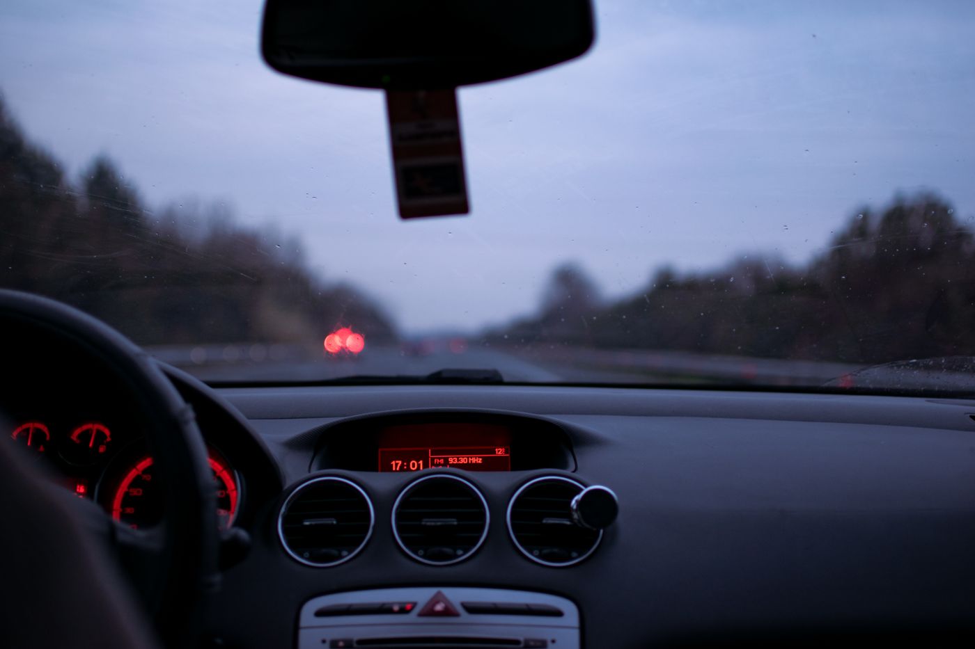A car dashboard glowing red at dusk, seen from the driver's seat, with a highway ahead through the windshield