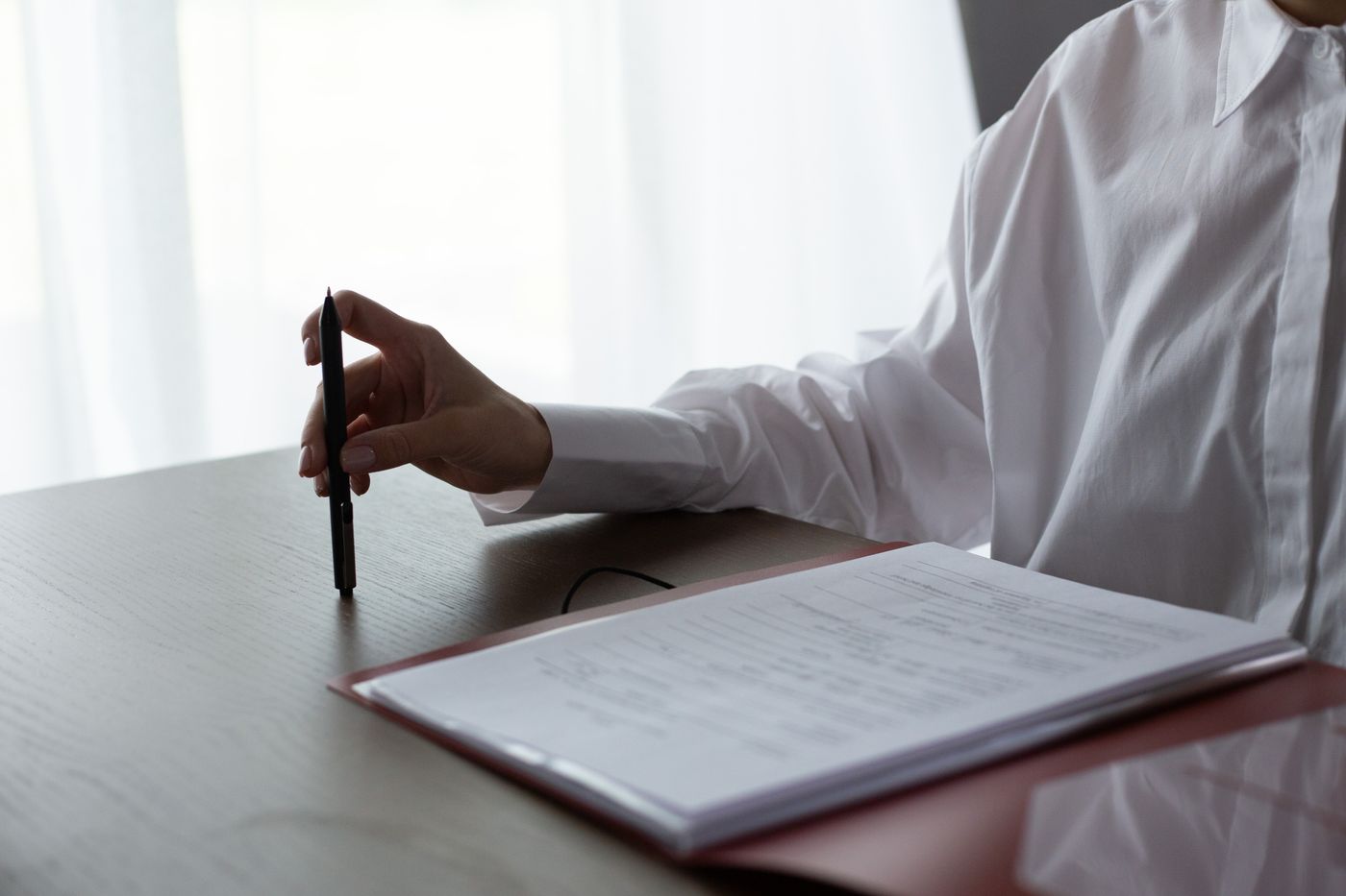 A person in a white shirt holding a pen beside a folder of papers on a wooden table