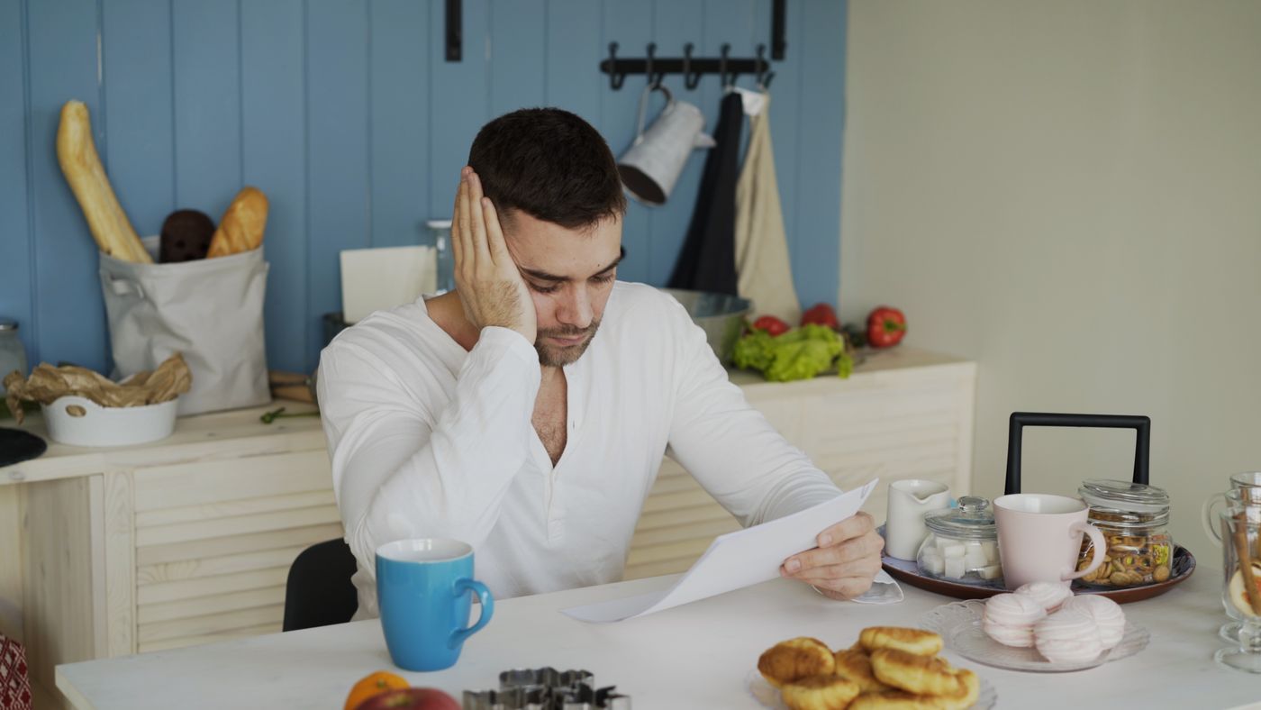 A worried man in a white shirt reading a sheet of paper at a kitchen table with coffee mugs and pastries